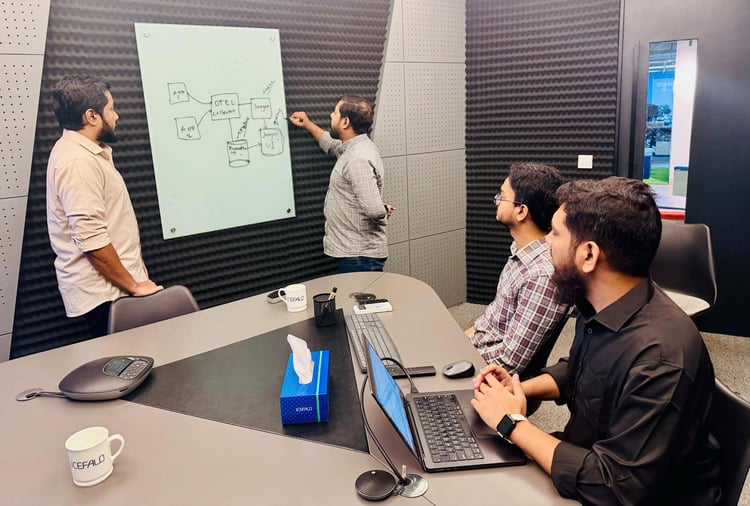 Fire menn har et teknisk møte i et møterom med lyddempende vegger. Én person står ved en whiteboard og tegner et diagram, mens de andre sitter rundt bordet og følger med. På bordet ligger bærbare PC-er, kaffekopper med Cefalo-logo og en eske med papirlommetørklær.