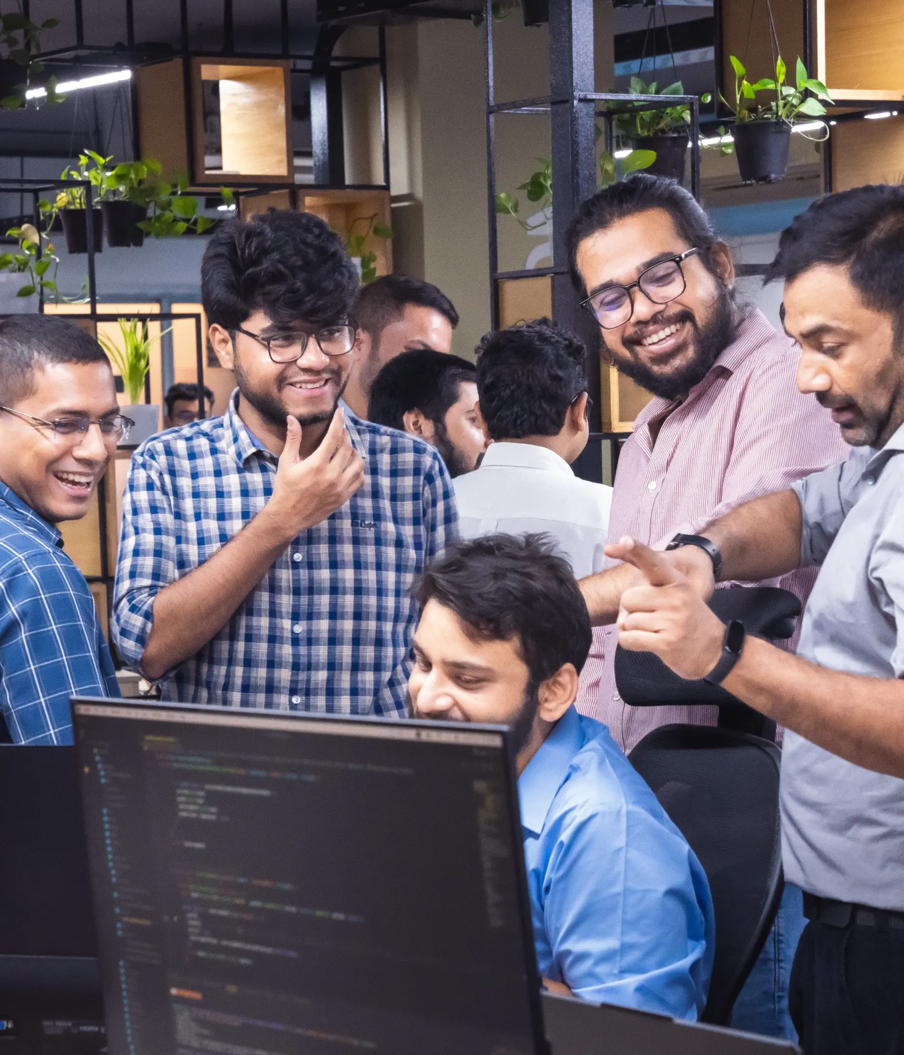 A group of developers discussing and laughing together around a computer screen in an open office space with shelves and plants in the background